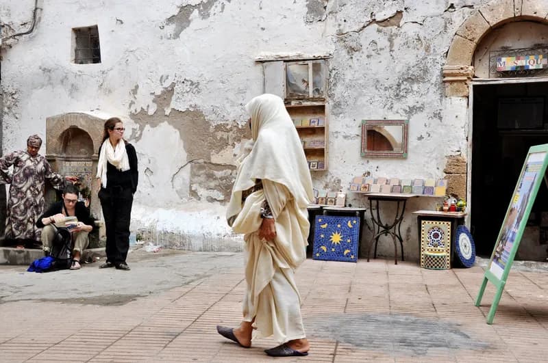 Une femme en haïk blanc traverse une placette du Mellah, devant des murs blanchis écaillés et un étal de carreaux peints