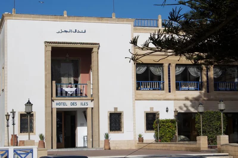 Façade blanche de l’Hôtel des Îles, avec son enseigne au-dessus de l’entrée et ses balcons à rambarde bleue