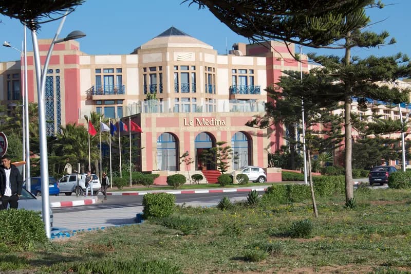 Façade rose et crème du Médina Essaouira Thalassa Sea & Spa, son enseigne au-dessus des arcades de l’entrée et des drapeaux