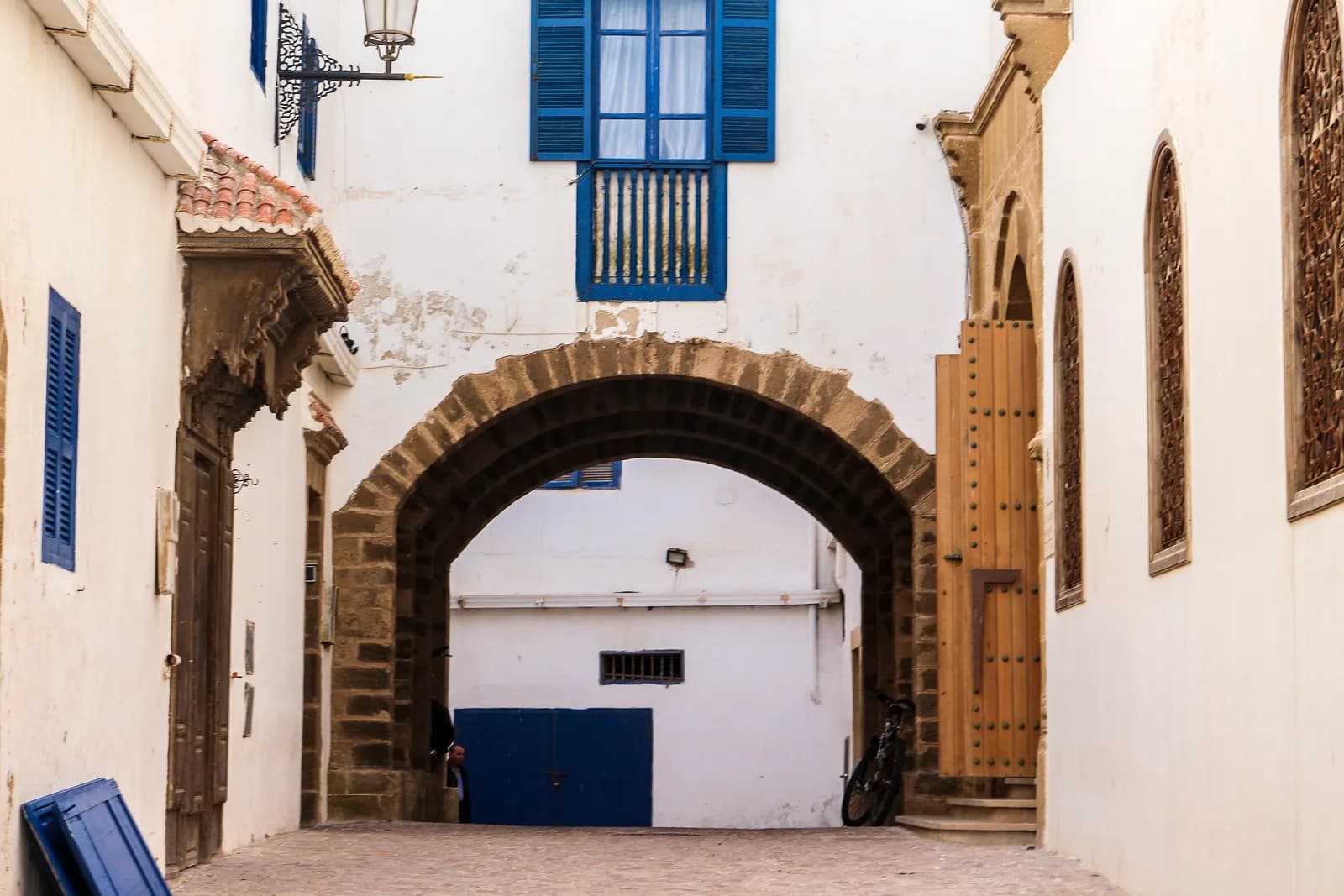 Ruelle pavée de la médina d’Essaouira passant sous une arche en pierre, murs blanchis, volets et portes bleus