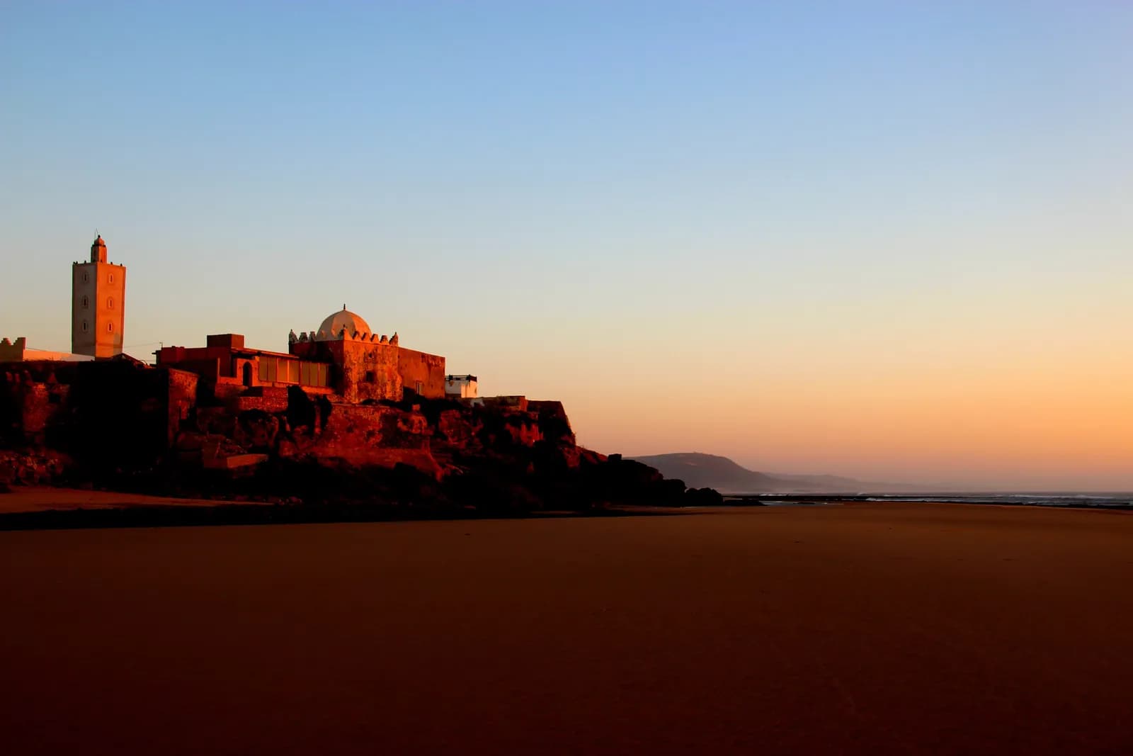 Minaret et coupole de Moulay Bouzerktoune sur leur promontoire rocheux, dans la lumière orangée d’un soleil bas, plage au premier plan