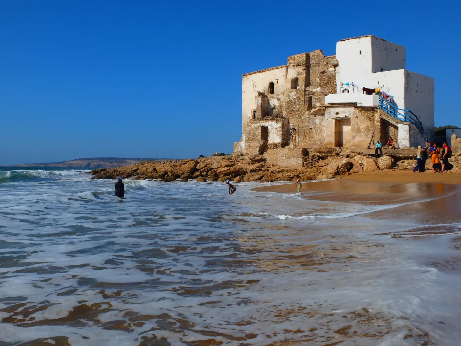Bâtisse blanchie en partie ruinée sur les rochers de Sidi Kaouki, baigneurs dans les vagues et sur le sable