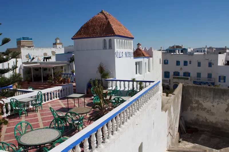 Toit-terrasse de la Villa Maroc, tables en mosaïque et chaises vertes au pied d’une tour blanche portant le nom de l’hôtel