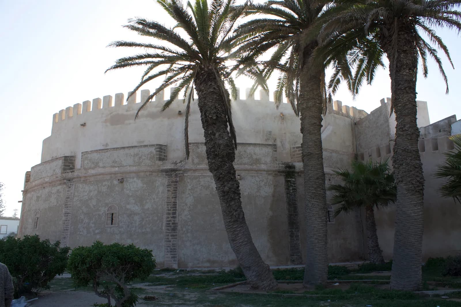 Grand bastion rond et crénelé du Borj Bab Marrakech, à contre-jour derrière des palmiers, dans un jardin