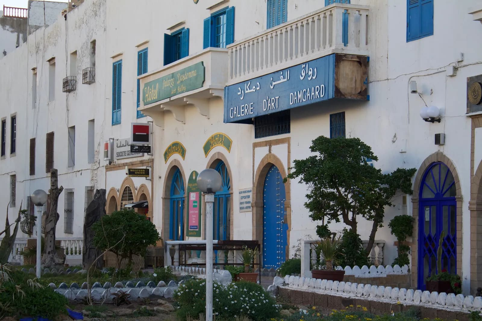 Enseigne bleue de la galerie Damgaard au-dessus d’une porte bleue en arc, façades blanches de l’avenue Oqba Ibn Nafiaa