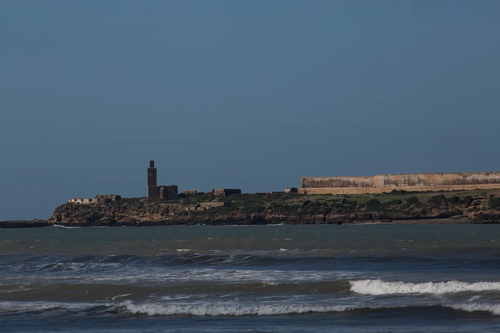 L’île de Mogador vue depuis la baie, une tour carrée et un long mur fortifié au-dessus des rochers, vagues au premier plan