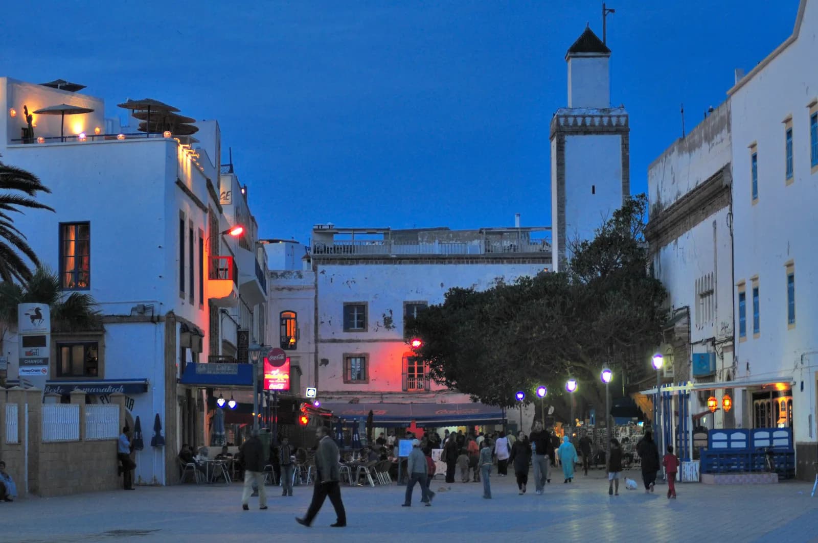 La place Moulay Hassan à la tombée de la nuit, terrasses de café éclairées, passants et minaret blanc au fond