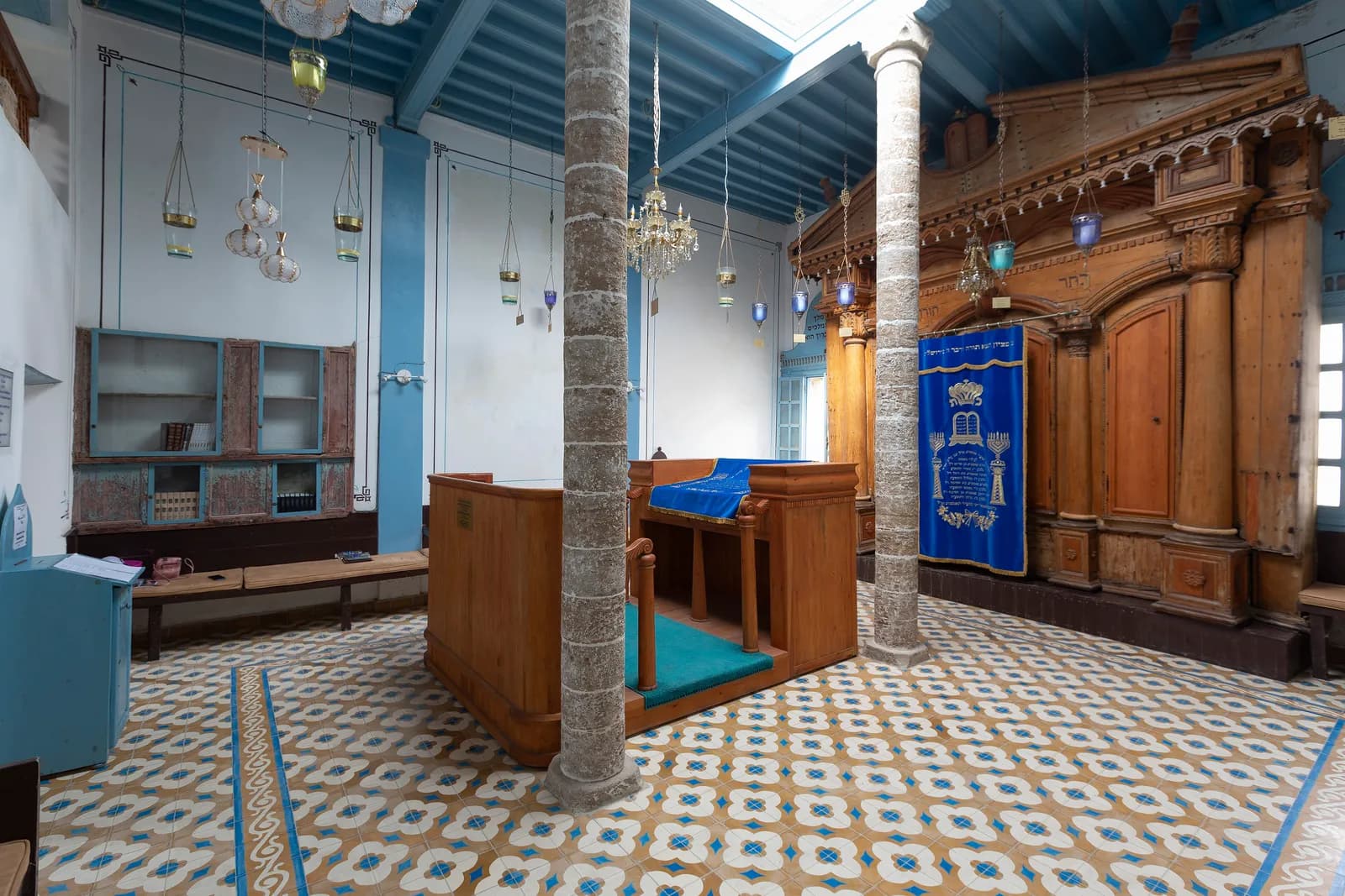 Salle de la synagogue Slat Lkahal : arche sainte en bois sculpté, rideau bleu brodé, tébah en bois, colonnes de pierre et sol en carreaux de ciment